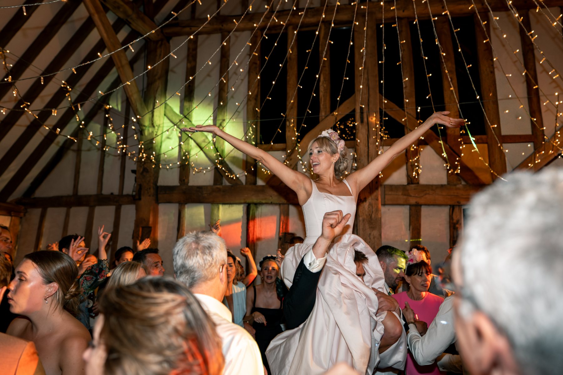 Bride lifted above a barn dancefloor with her arms outstretched under strings of fairy lights