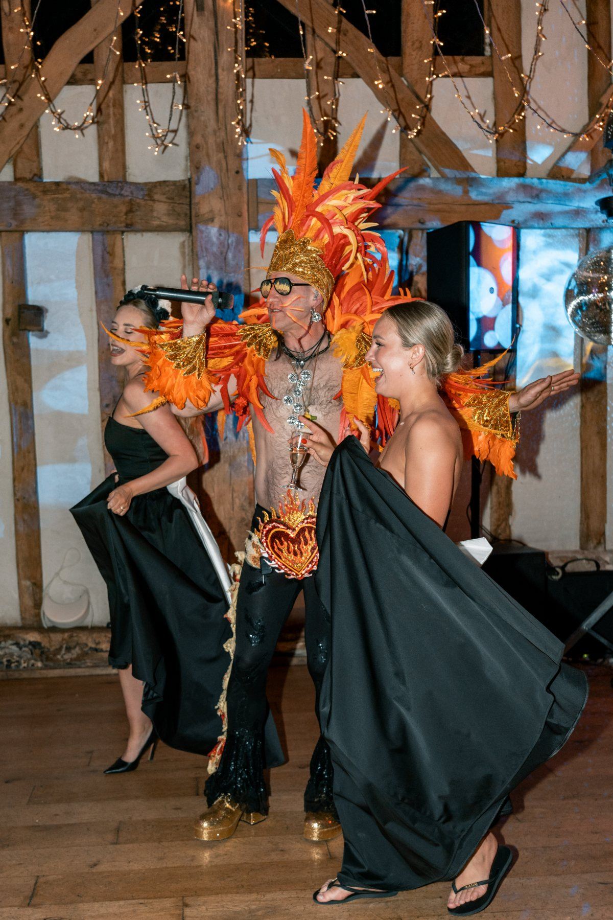 Two guests swinging their gowns either side of the frontman as he sings in a timber framed barn
