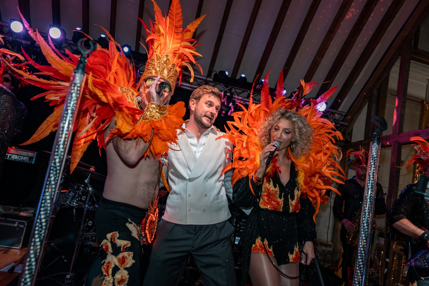 Groom in a white waistcoat pulled onstage between two performers in orange feather headdresses