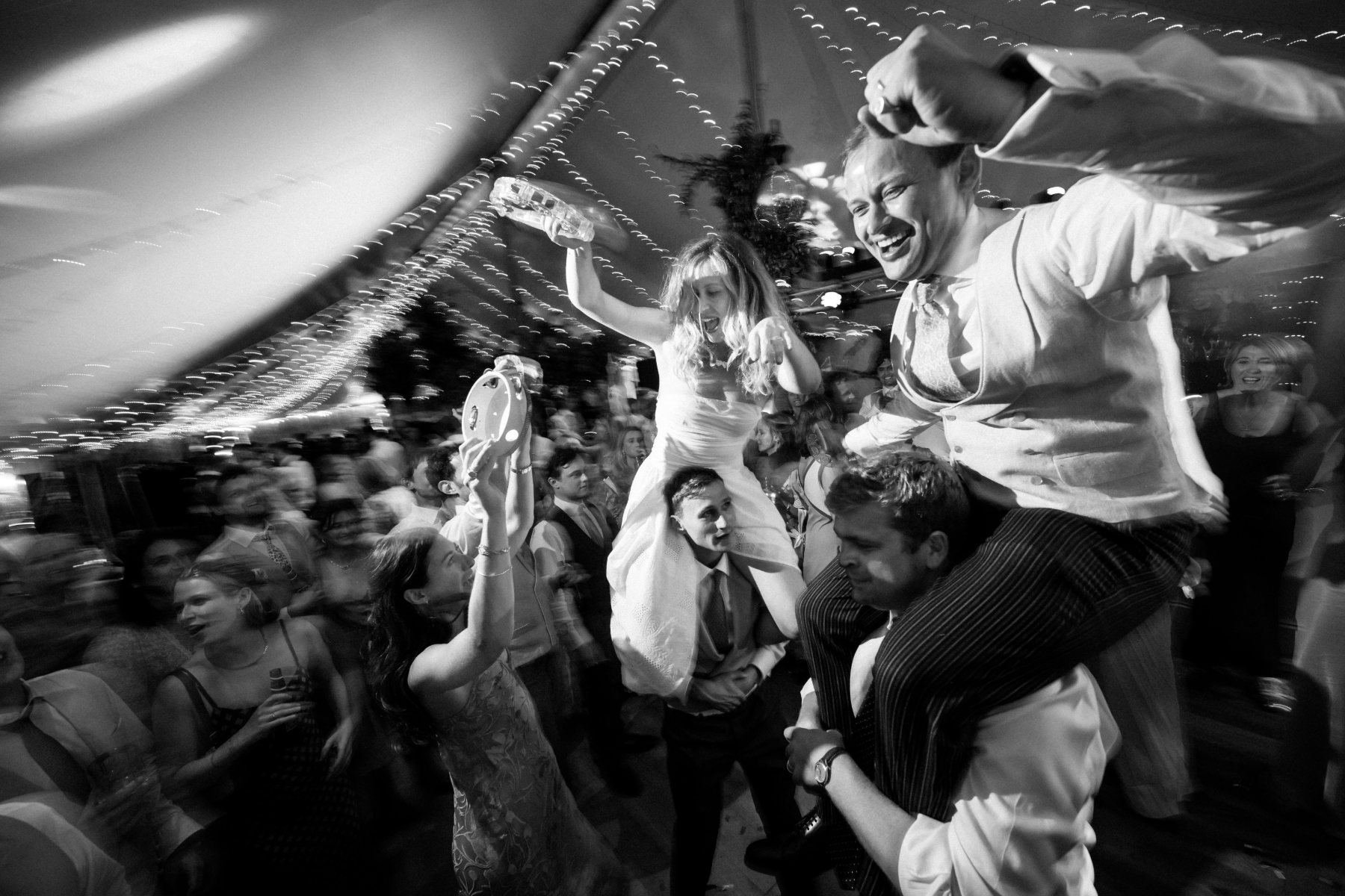 Bride and groom both lifted on shoulders on a packed dancefloor, a guest shaking a tambourine beside them