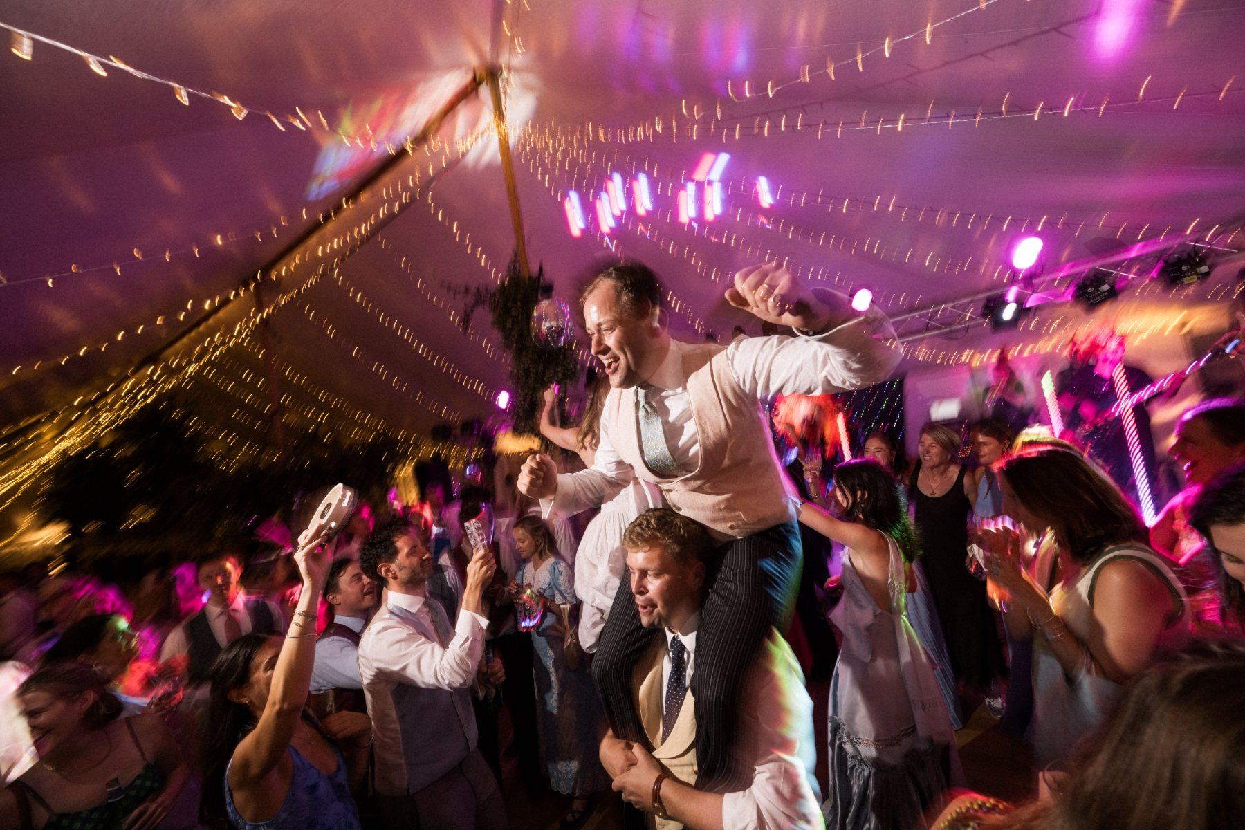 Groom on a guest's shoulders with his fist in the air under the marquee lights