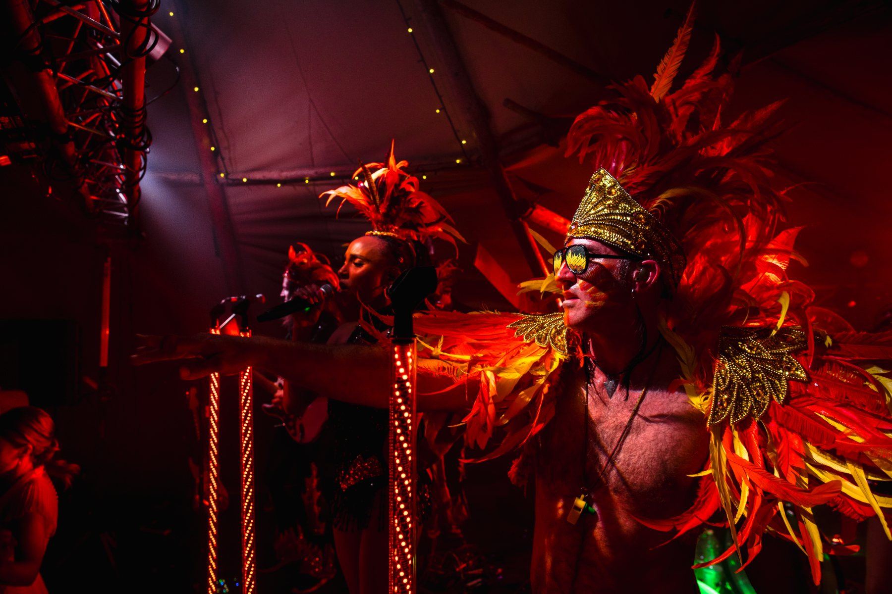 Uncle Funk's frontman and vocalist mid song under deep red light, feather headdresses glowing beside LED mic stands